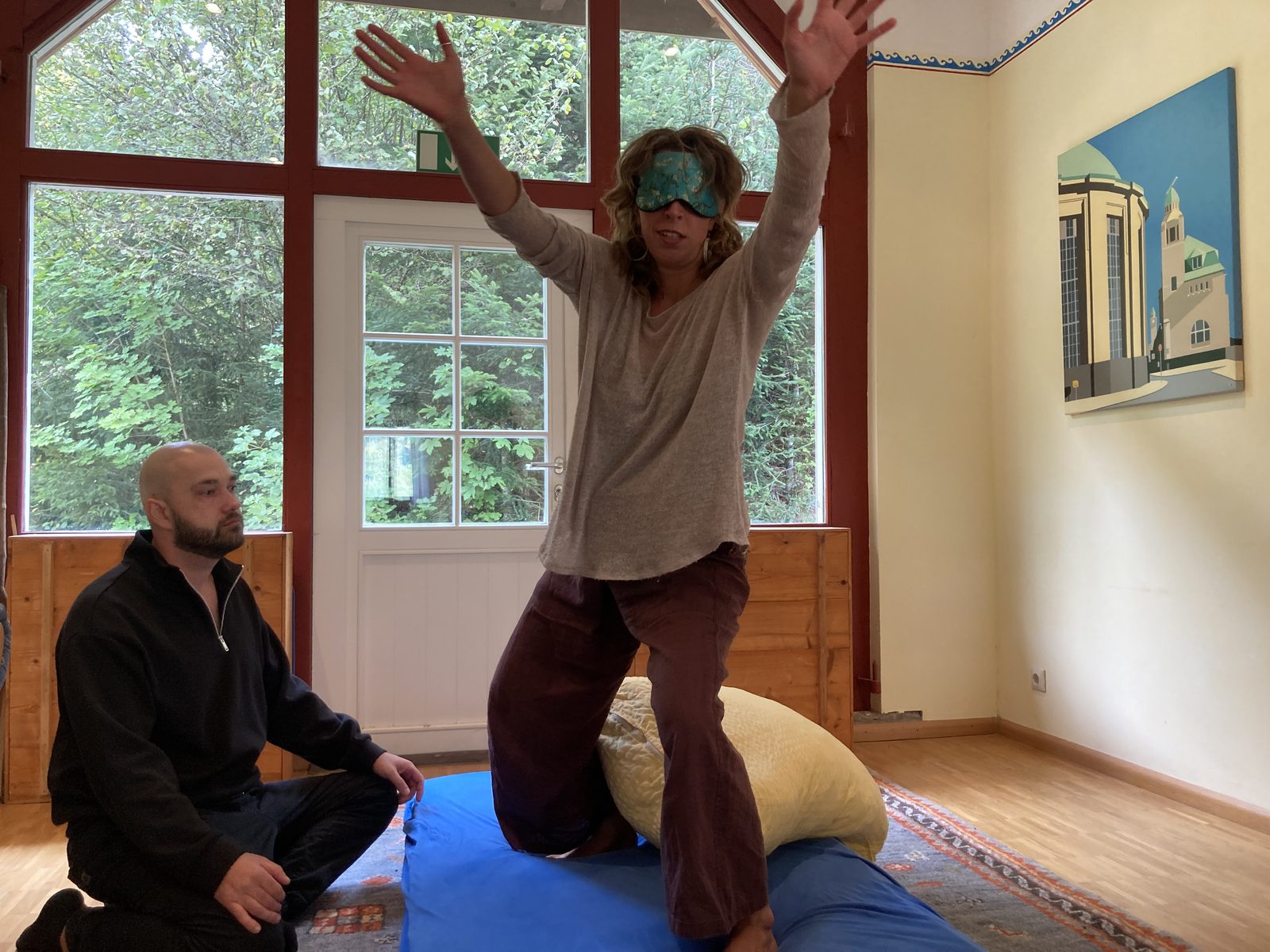 A blindfolded participant moving with arms raised, watched by her sitter