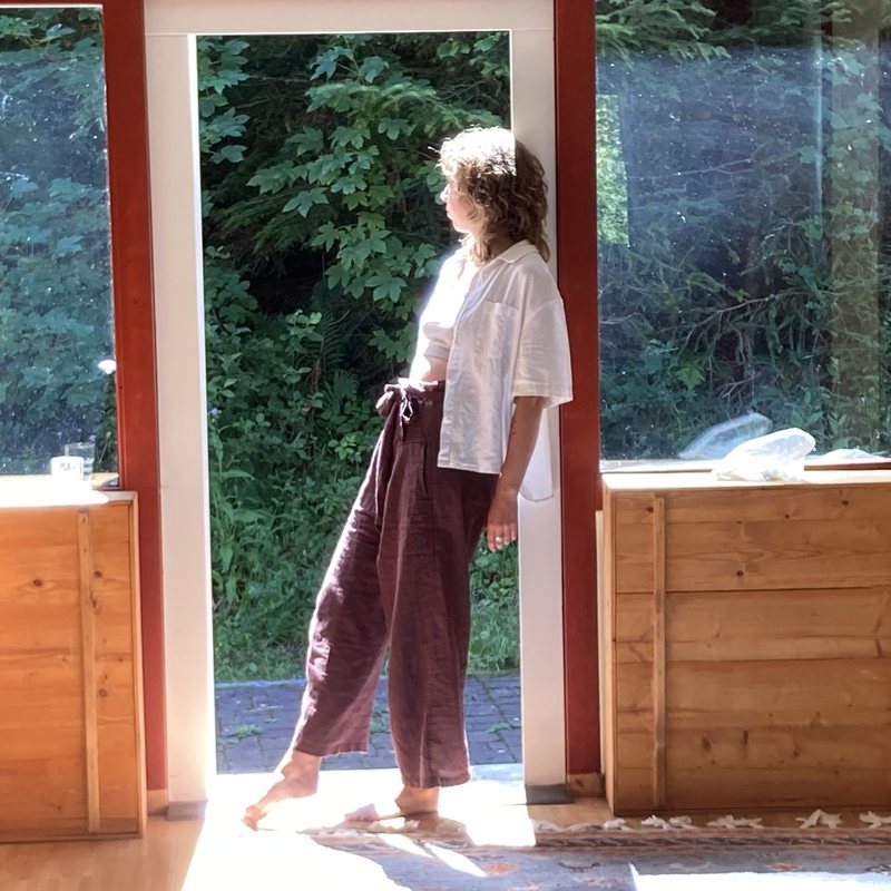 A barefoot woman standing in a sunlit open doorway, looking out towards the green woodland beyond